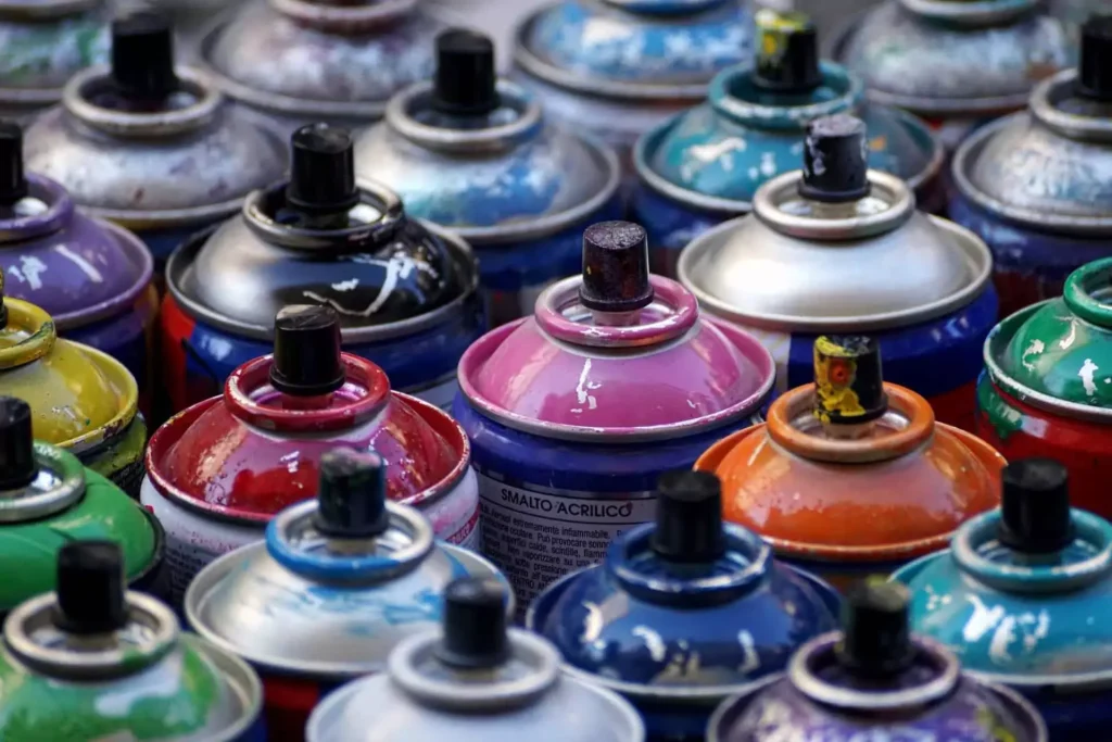A close-up view of numerous spray paint cans arranged tightly together, showing a variety of bright colors including pink, red, blue, green, orange, yellow, and black. The cans are well-used, with paint splatters and worn nozzles, and the word 'SMALTO ACRILICO' is visible on one can’s label.