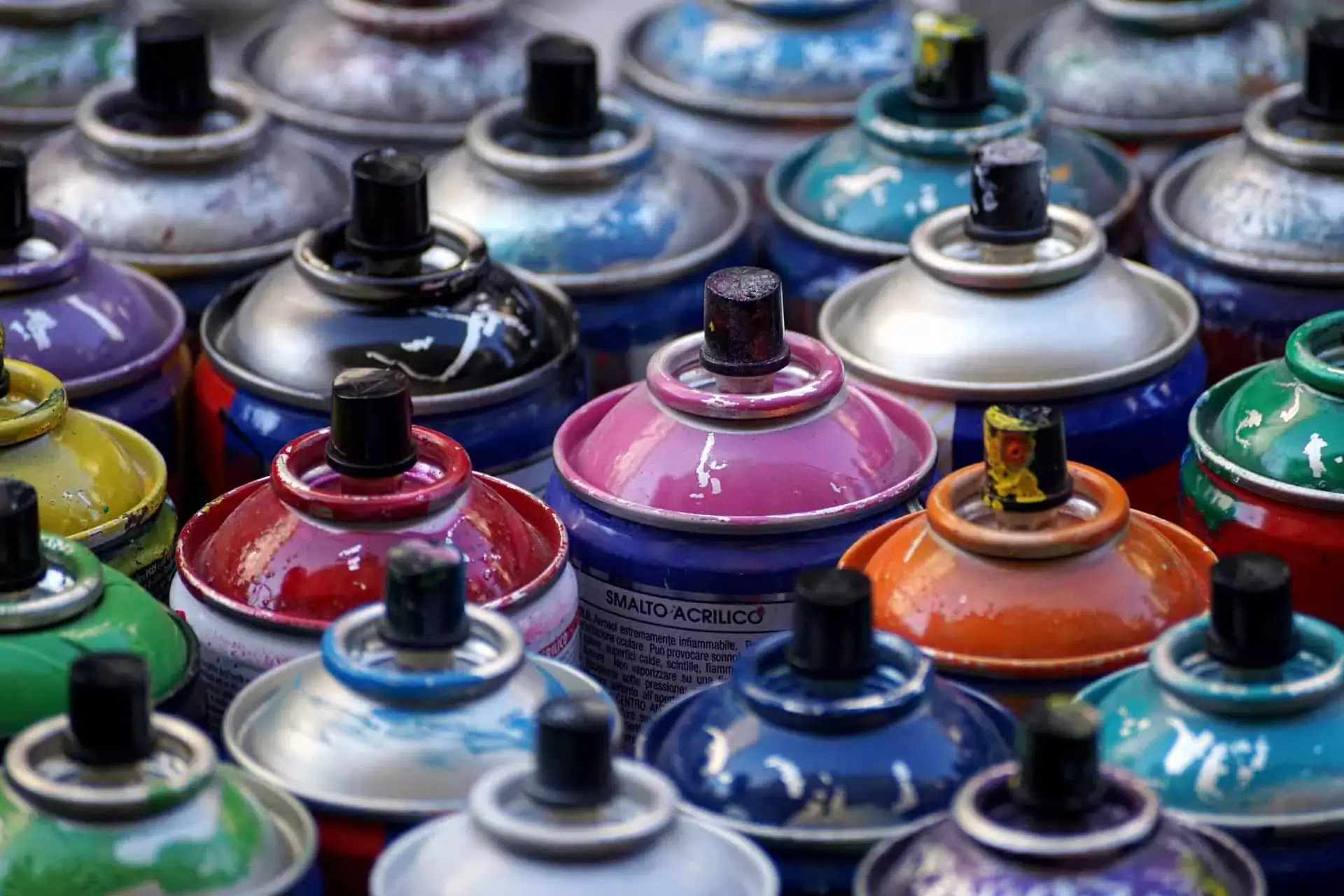 A close-up view of numerous spray paint cans arranged tightly together, showing a variety of bright colors including pink, red, blue, green, orange, yellow, and black. The cans are well-used, with paint splatters and worn nozzles, and the word 'SMALTO ACRILICO' is visible on one can’s label.