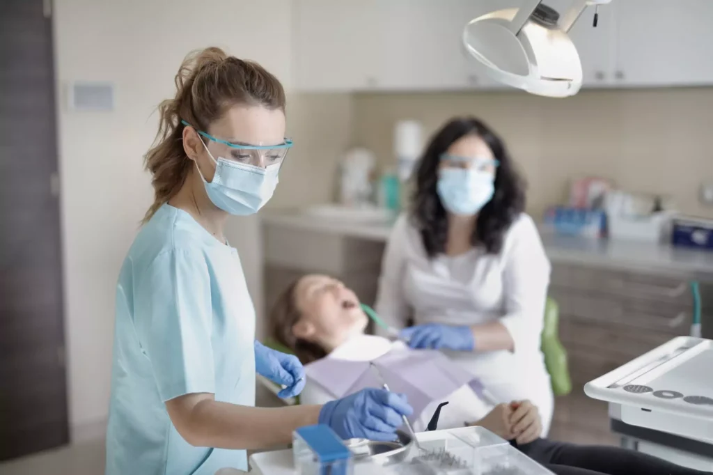 A white female dental hygienist, wearing a light blue scrub top, a medical mask, and clear safety glasses, stands over a young patient in a dental chair. She is focused on her work, holding a dental tool with blue-gloved hands. The patient, a child, is lying back with their mouth open. A female dentist, also wearing a medical mask and glasses, is in the background, assisting with the procedure. The scene takes place in a clean, brightly lit dental office.