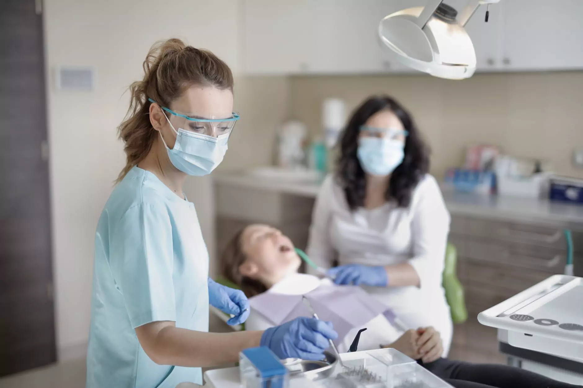A white female dental hygienist, wearing a light blue scrub top, a medical mask, and clear safety glasses, stands over a young patient in a dental chair. She is focused on her work, holding a dental tool with blue-gloved hands. The patient, a child, is lying back with their mouth open. A female dentist, also wearing a medical mask and glasses, is in the background, assisting with the procedure. The scene takes place in a clean, brightly lit dental office.