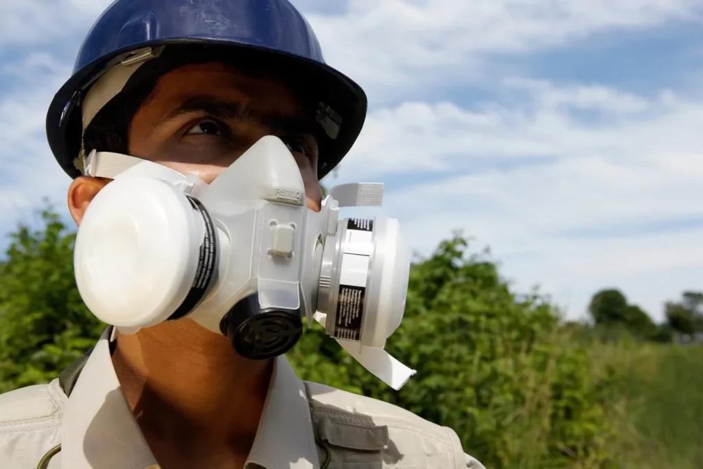 A construction worker or technician wearing a blue hard hat and a white respirator mask looks up and to the side. The sky is visible in the background, with greenery at the bottom of the frame.