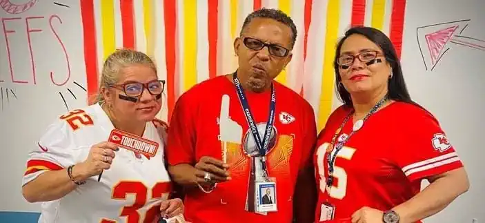 Three people, two women and one man, are posing in Kansas City Chiefs-themed attire. The woman on the left, who is white, is wearing a white Chiefs jersey with the number ‘32’ on it. She has black face paint under her eyes and is holding a ‘TOUCHDOWN’ sign. The man in the middle, who is Black, is wearing a red Chiefs t-shirt, glasses, and a lanyard with an ID badge. He has a playful, puckered-lip expression. The woman on the right, who is Hispanic, is wearing a red Chiefs jersey with the number ‘15’ and has black face paint under her eyes. The background is decorated with red and yellow streamers and a hand-drawn sign that says ‘CHIEFS’.