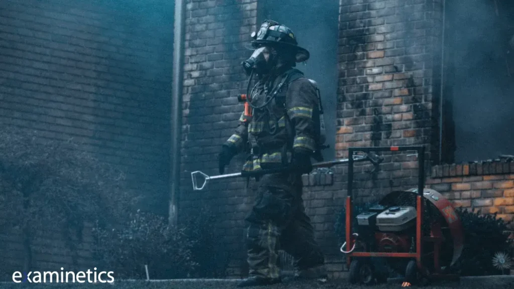 A firefighter in full protective gear, including a helmet, face mask, and oxygen tank, walks past a brick building with smoke billowing in the background. The firefighter carries a long metal tool and is next to a red portable fan or ventilation unit on wheels. The scene appears to be during active firefighting operations.