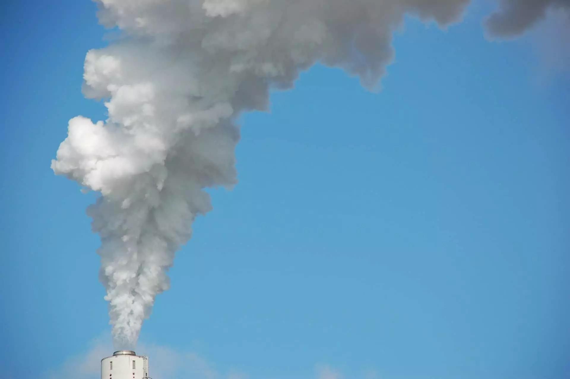 A large industrial smokestack releasing a thick plume of white smoke or steam into a clear blue sky. The emission billows upward and spreads out, creating dense, cloud-like formations against the otherwise cloudless background.