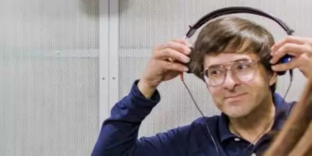 A close-up shot of a white man with glasses and short brown hair, wearing a dark blue polo shirt. He is smiling slightly while adjusting a pair of large, black headphones with blue earpieces on his head, as if he is preparing for a hearing test. The background is a grey, textured wall, likely inside a sound booth.