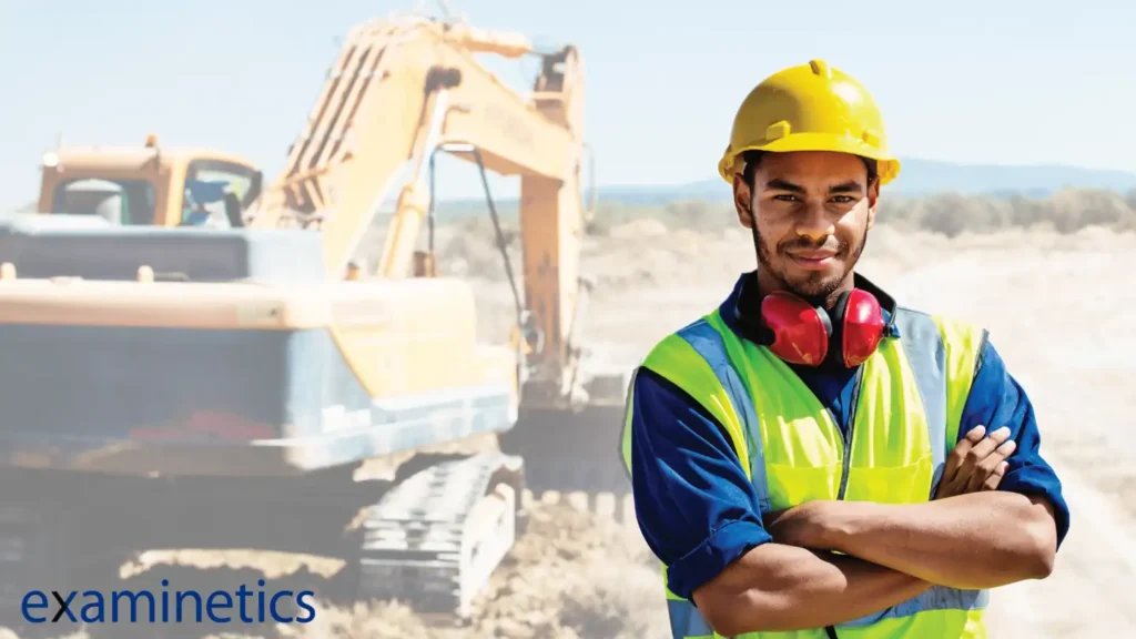 A male construction worker of color, wearing a yellow hard hat and a high-visibility yellow-green vest over a blue work shirt, stands with his arms crossed and smiles confidently at the camera. He has large red earmuffs for hearing protection resting around his neck. In the blurred background, there is a large yellow excavator on a dirt construction site under a bright, sunny sky. The bottom left corner of the image has a logo that reads ‘examinetics’ in blue text.