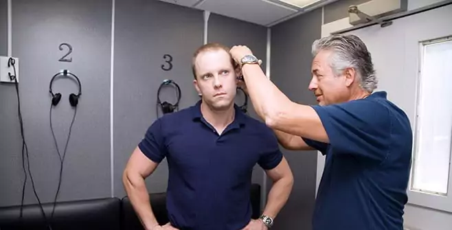 A man in a navy blue polo shirt stands with his arms on his hips, looking straight ahead with a serious expression, while another man with gray hair, also wearing a navy polo shirt, adjusts or examines something near his ear. The setting appears to be a small room with numbered headphone stations mounted on the wall behind them.