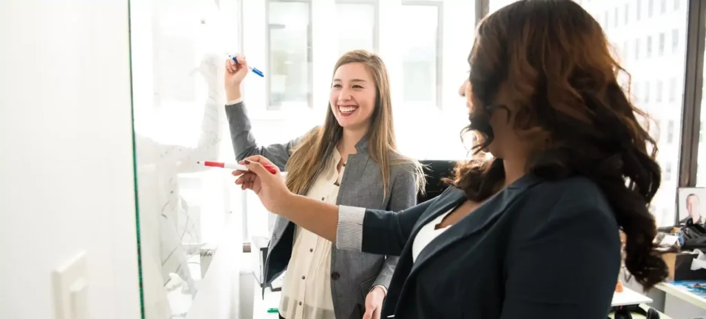 Two professional women are collaborating at a whiteboard in a bright, modern office. The woman on the left, a white woman with long brown hair, is laughing as she holds a blue marker and writes on the board. The woman on the right, a Black woman with long dark hair, holds a red marker and is gesturing toward the board. Both women are wearing business jackets, and their interaction appears friendly and collaborative. The background shows a window with a view of other buildings and a glimpse of a third person.