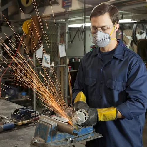 A focused white male worker is using a hand-held grinder to cut a metal pipe secured in a vise on a workbench. He is wearing a dark blue long-sleeved work shirt, a pleated respirator mask, clear safety glasses, yellow earplugs, and black and yellow work gloves. Sparks are flying from the grinder, creating a bright shower of light. The background is a cluttered workshop with various tools, equipment, and a yellow hard hat visible.