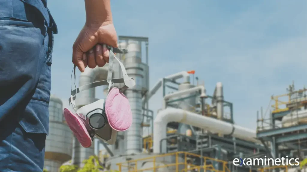 Close-up of a worker’s hand holding a respirator mask with pink filters, standing in front of an industrial facility with large metal pipes, tanks, and structures under a clear blue sky. The worker is wearing dark blue work pants, and the scene conveys a focus on safety in an industrial environment.