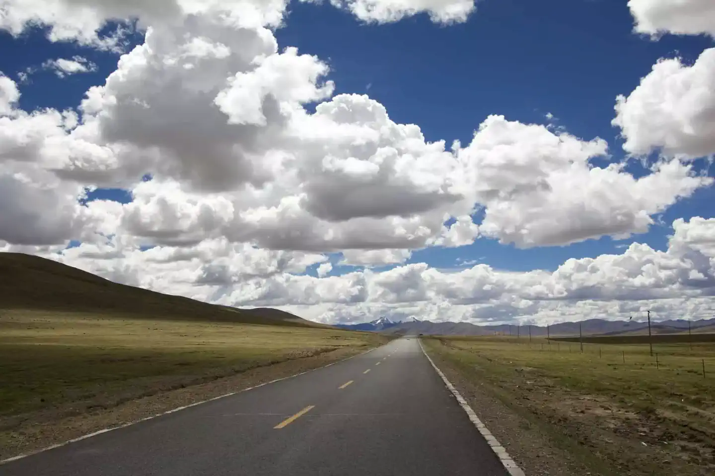 A straight, paved two-lane road with a broken yellow line runs through a vast, grassy plain. The road leads toward the distant horizon where mountains are visible under a bright blue sky filled with large, puffy white clouds. The landscape on either side of the road is a rolling, grassy, brownish-green prairie.