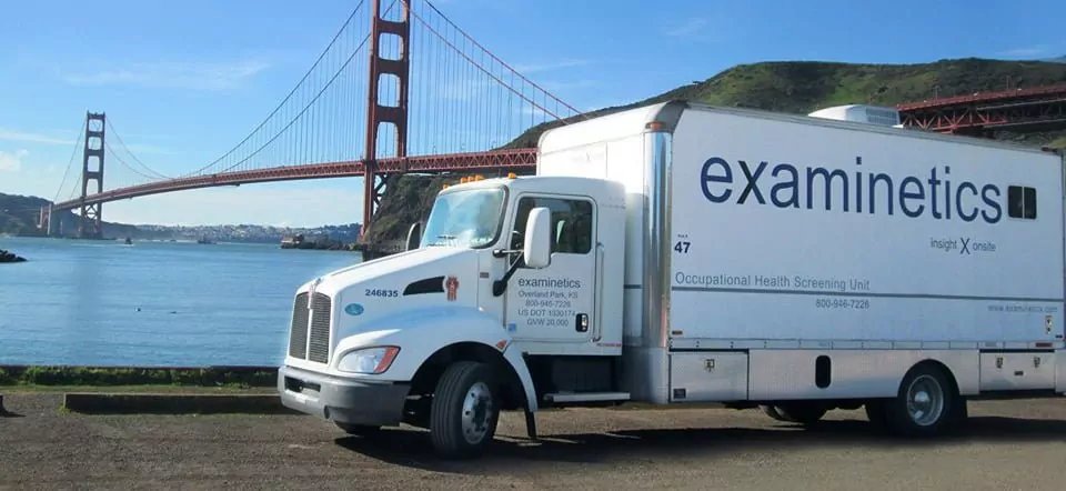 A large white truck labeled ‘examinetics’ with the tagline ‘insight X onsite’ is parked with the Golden Gate Bridge in San Francisco visible in the background. The bridge is a reddish-orange color, spanning a body of blue water. The truck has ‘Occupational Health Screening Unit’ printed on its side along with a phone number and website. The sky is bright blue with a few scattered clouds, and green hills are visible behind the bridge.
