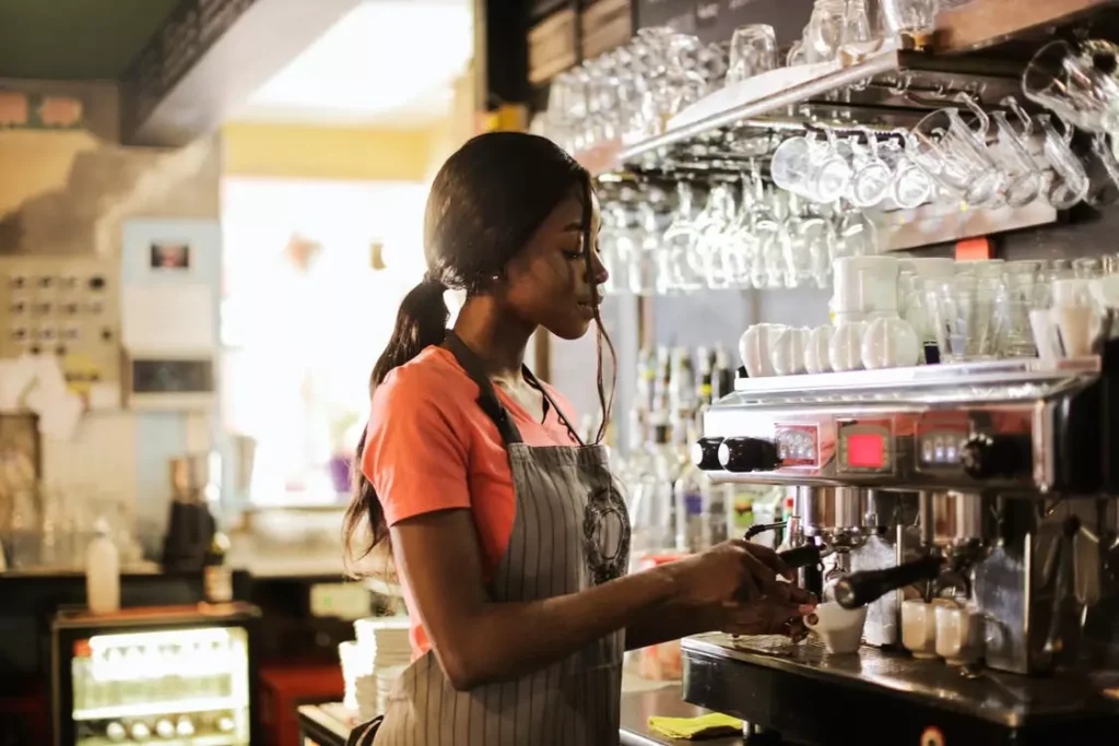 A focused Black woman with her hair in a ponytail, wearing an orange t-shirt and a grey pinstripe apron, operates a professional espresso machine in a dimly lit cafe. She holds a small white cup under the machine’s nozzle with her right hand while her left hand is on the machine’s lever. Above her, rows of clean, inverted glasses and mugs hang from a rack. The background is slightly blurred but shows a bright window and some shelving.
