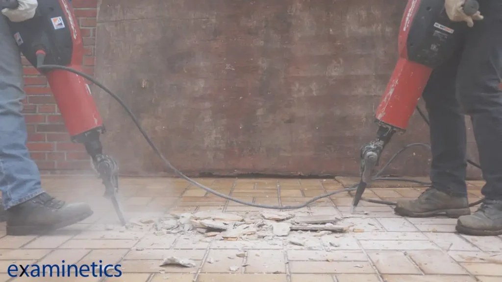 Two people are operating large red jackhammers, breaking up a tiled floor. The image focuses on their legs and the jackhammers. The person on the left is wearing blue jeans and work boots, and the person on the right is wearing dark pants and work boots. Dust and small pieces of debris are flying up from the floor as the jackhammers work. The background is a brick wall. A blue ‘examinetics’ logo is in the bottom left corner.