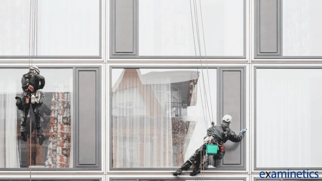 Two window washers, suspended by ropes, clean the exterior of a modern multi-story building. The window washer on the left, seen from behind, is wearing a black jacket and a white helmet. The window washer on the right is also wearing a black jacket and a white helmet, and is actively wiping a window. They are both harnessed and have various tools and buckets hanging from their belts. The building has a grid of large windows with grey frames. The windows reflect the surrounding buildings and a cloudy sky. The ‘examinetics’ logo is in the bottom right corner in white text.