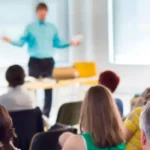 A panoramic view from behind an audience sitting in rows of chairs at a conference or training class. A man in a blue shirt is standing in the front, facing the audience, with his arms outstretched. He is blurred and appears to be giving a presentation. A large screen is visible on the left side of the frame. The audience's backs are visible, and they are all facing the speaker. The room is brightly lit with large windows in the background.