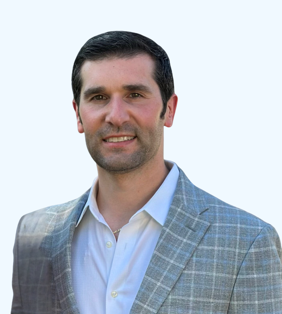 Portrait of a man with short dark hair and light stubble, wearing a light plaid blazer over a white button-down shirt, against a clean, light background.